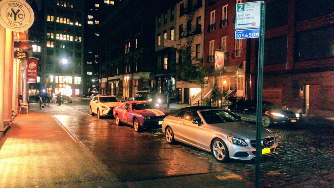 A wet cobblestone stretch of Bond Street in Manhattan at night, with parked cars and lit storefronts